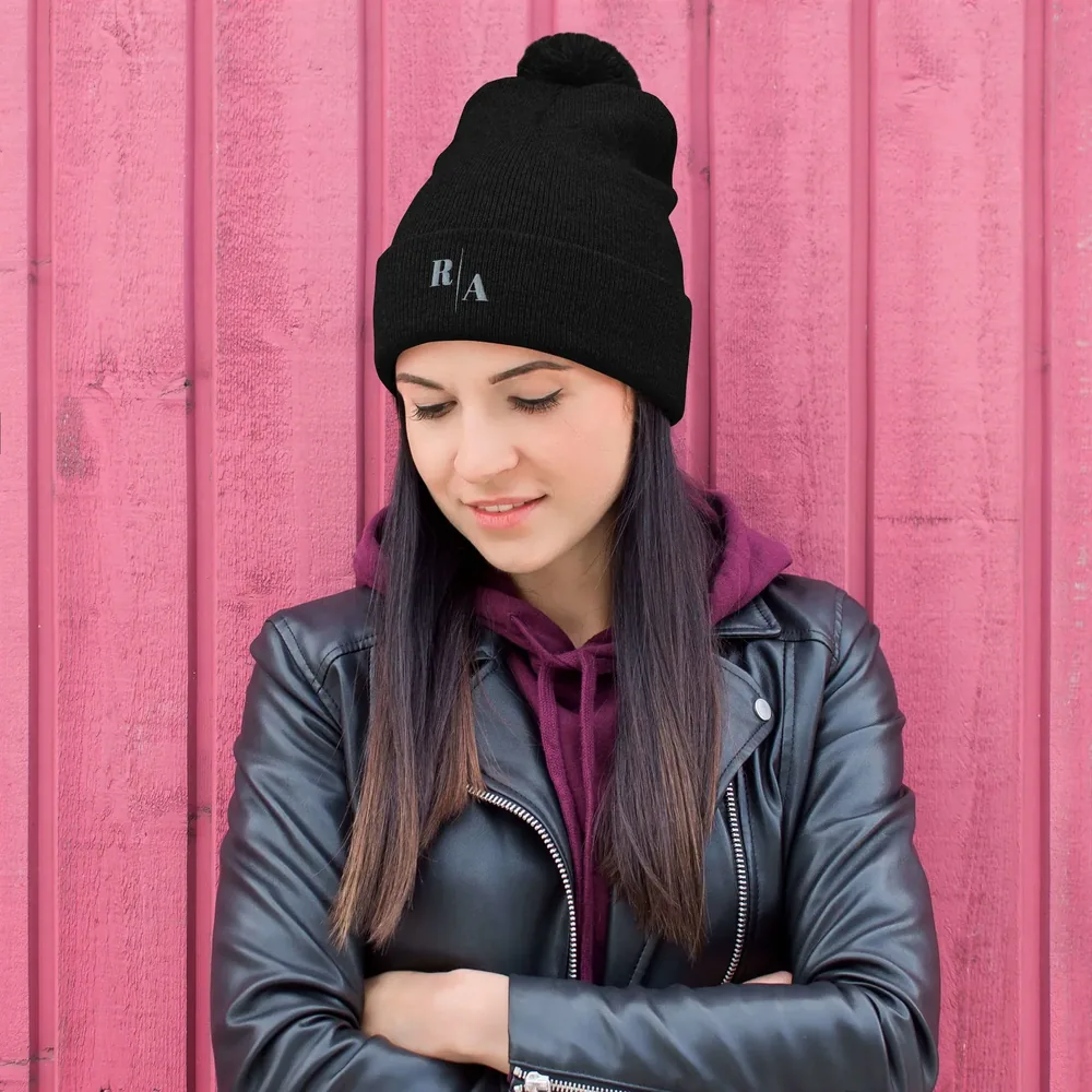 A woman wears the black Timeless Cap, a pom-pom beanie embroidered with the R|A mark, against a pink timber wall.